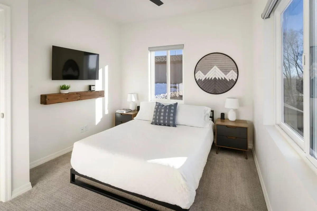A serene bedroom filled with natural light from a large window, featuring elegant wall art, a nightstand, and a comfortable bed. The window offers potential sunset or mountain views, creating an inviting atmosphere for relaxation and contemplation.