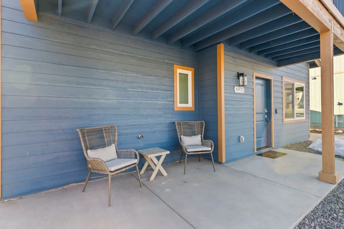 This image showcases a welcoming front porch with two wicker chairs and a small side table set against a blue wall. Morning sunlight streams onto the porch, creating an inviting spot for coffee or relaxed conversation.