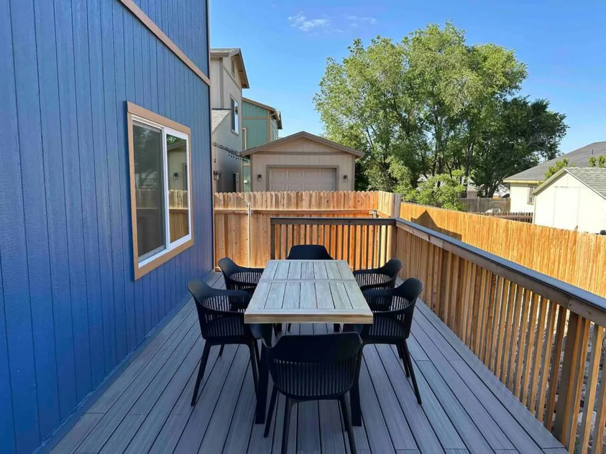 An inviting outdoor dining area with a table and chairs set beside the home's blue exterior wall and a fenced yard. The comfortable setup encourages group meals, sunset dinners, and relaxed mornings in nature.