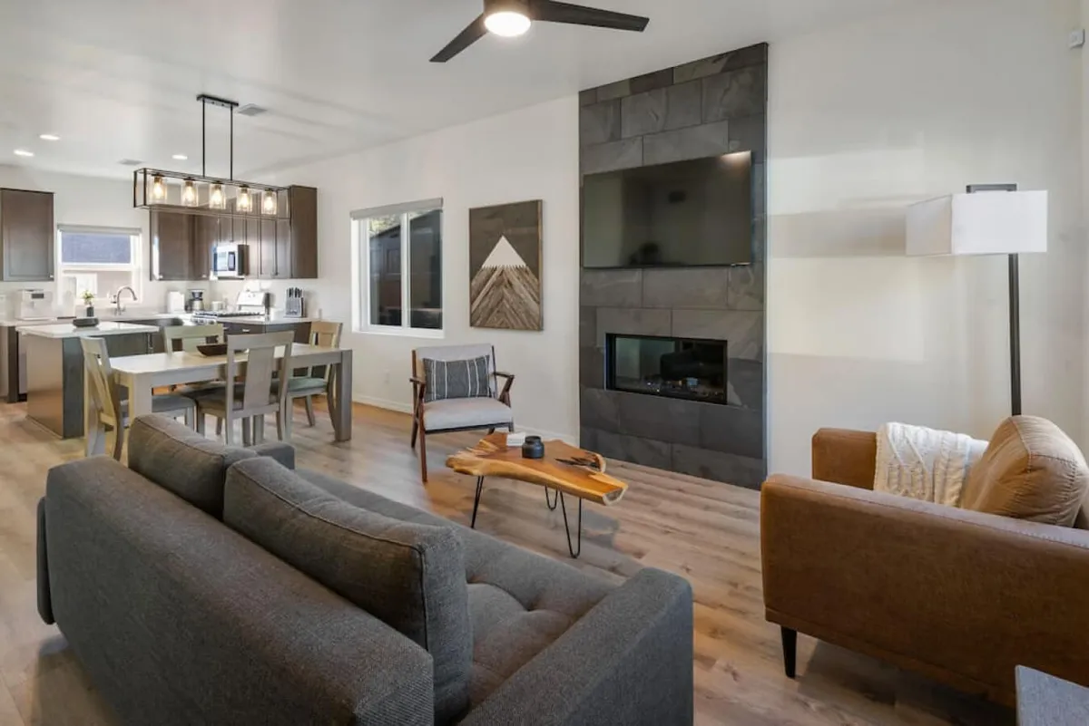 The image showcases a stylish, modern living and dining area with a cozy sofa, a chic dining table set for gathering, a warm fireplace as the focal point, and tasteful artwork adorning the walls. Abundant natural light fills the space, highlighting the clean lines and inviting atmosphere.
