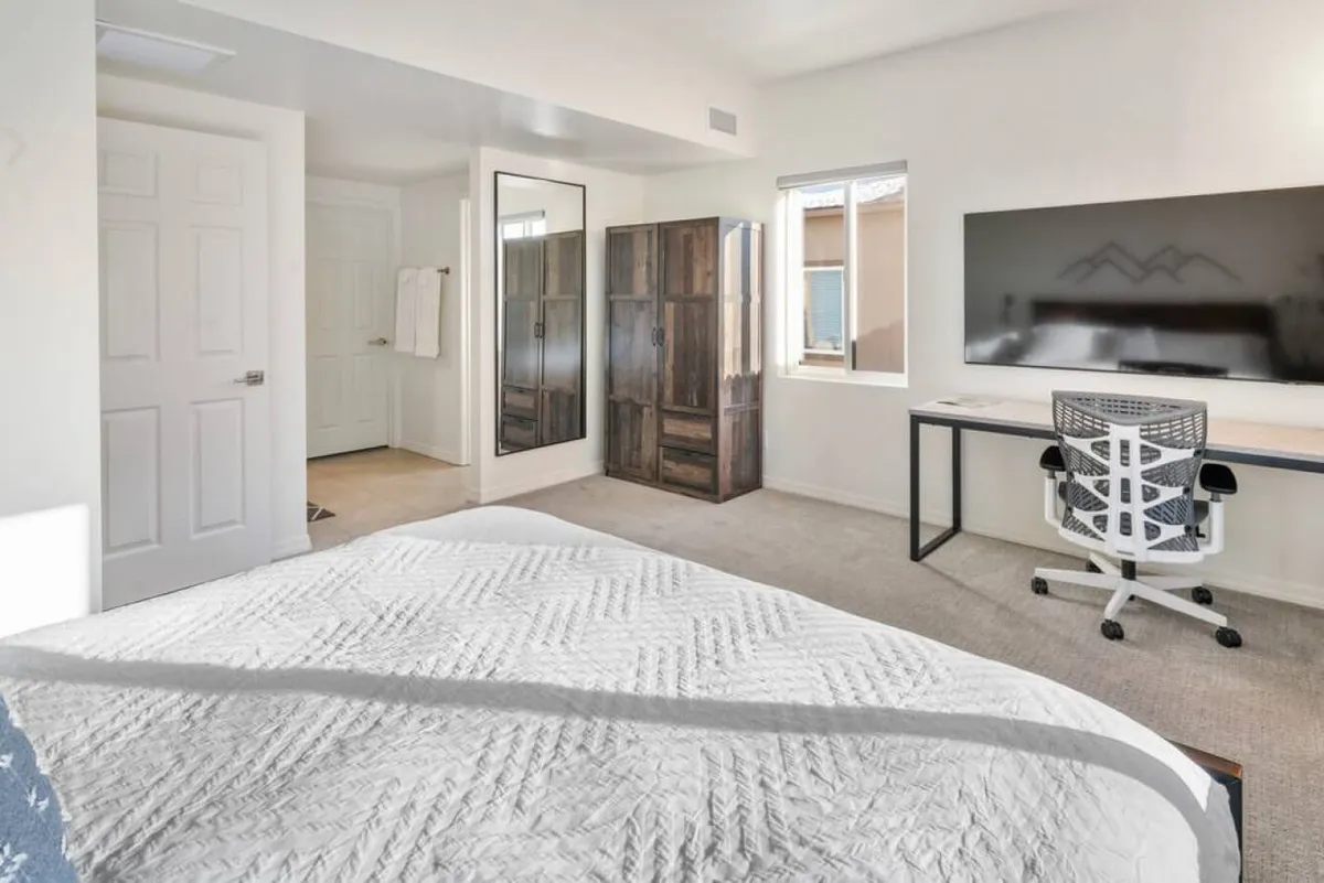 A bright and organized bedroom featuring a neatly made bed with textured white bedding, a large flat-screen TV on the wall, and a modern desk with an ergonomic chair beside a window letting in natural light. The space feels secure and orderly, offering an inviting environment for relaxation and focus.