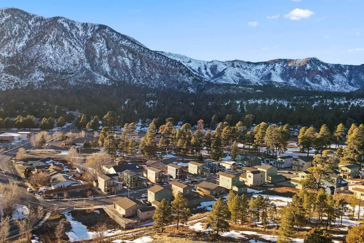 This image captures a breathtaking view of snow-capped mountains rising over a peaceful neighborhood lined with pine trees. The foreground features a quiet residential street with homes nestled into the landscape, while the background is dominated by the dramatic slopes of Mount Elden and its surrounding ranges. The snowy scenery hints at year-round exploration and the natural beauty awaiting just steps from the front door.