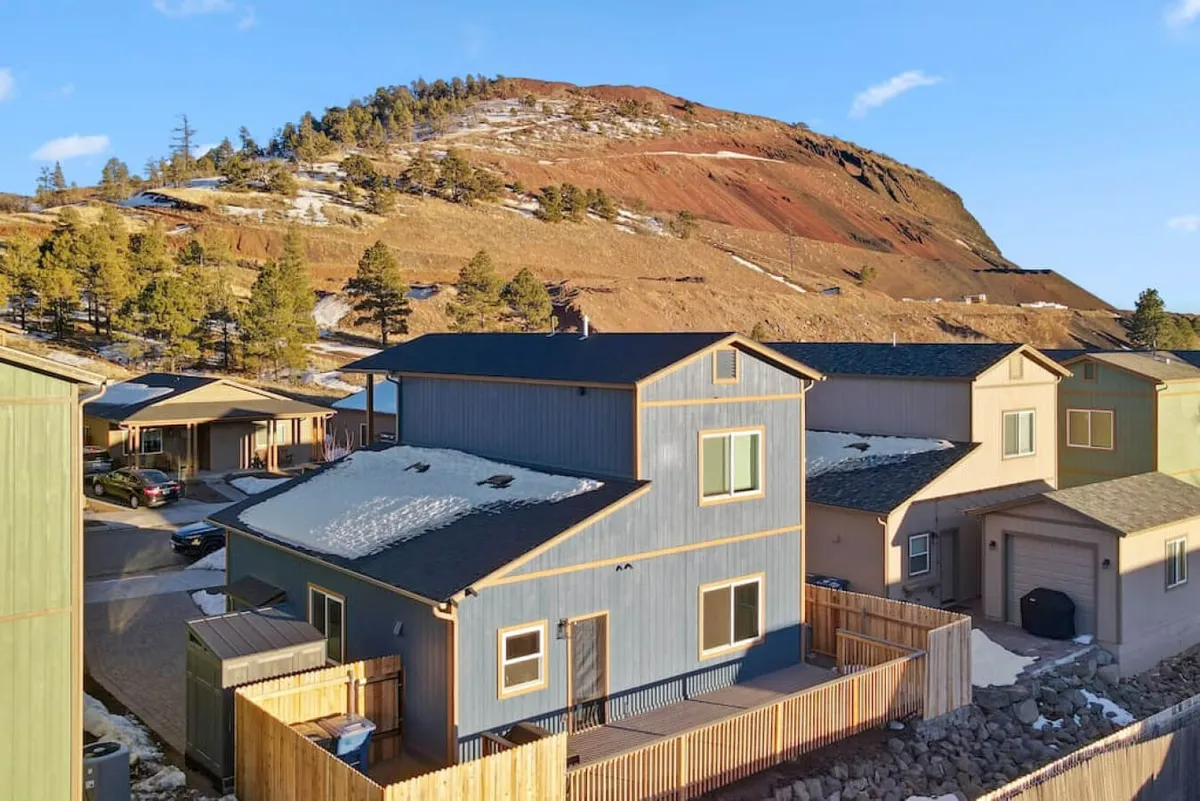 The image shows a charming blue house nestled against a stunning mountain backdrop, surrounded by serene natural landscaping. The scene evokes feelings of peace and adventure, with plenty of open space for outdoor fun and relaxation.