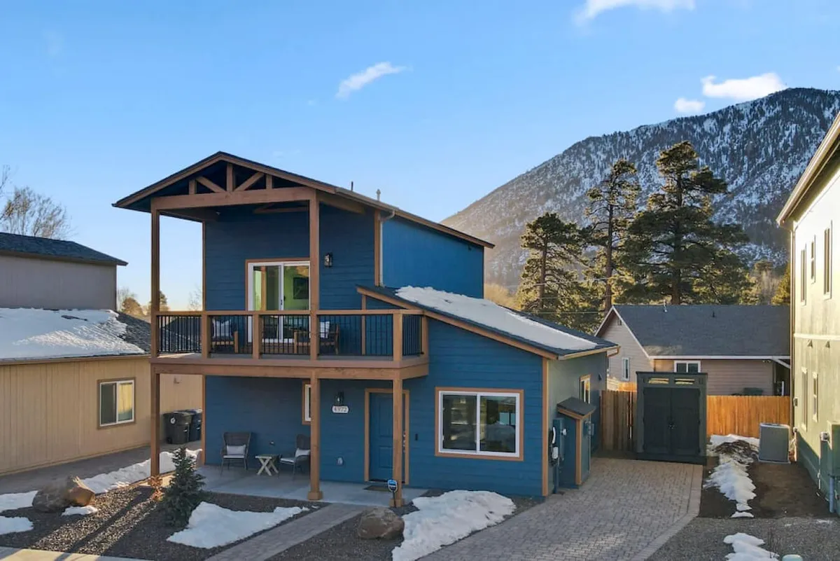 The exterior of the property showcases a welcoming blue facade with a balcony, set against a backdrop of serene mountains and open skies. The entryway is prominent, suggesting secure access and peaceful surroundings in a modern neighborhood.