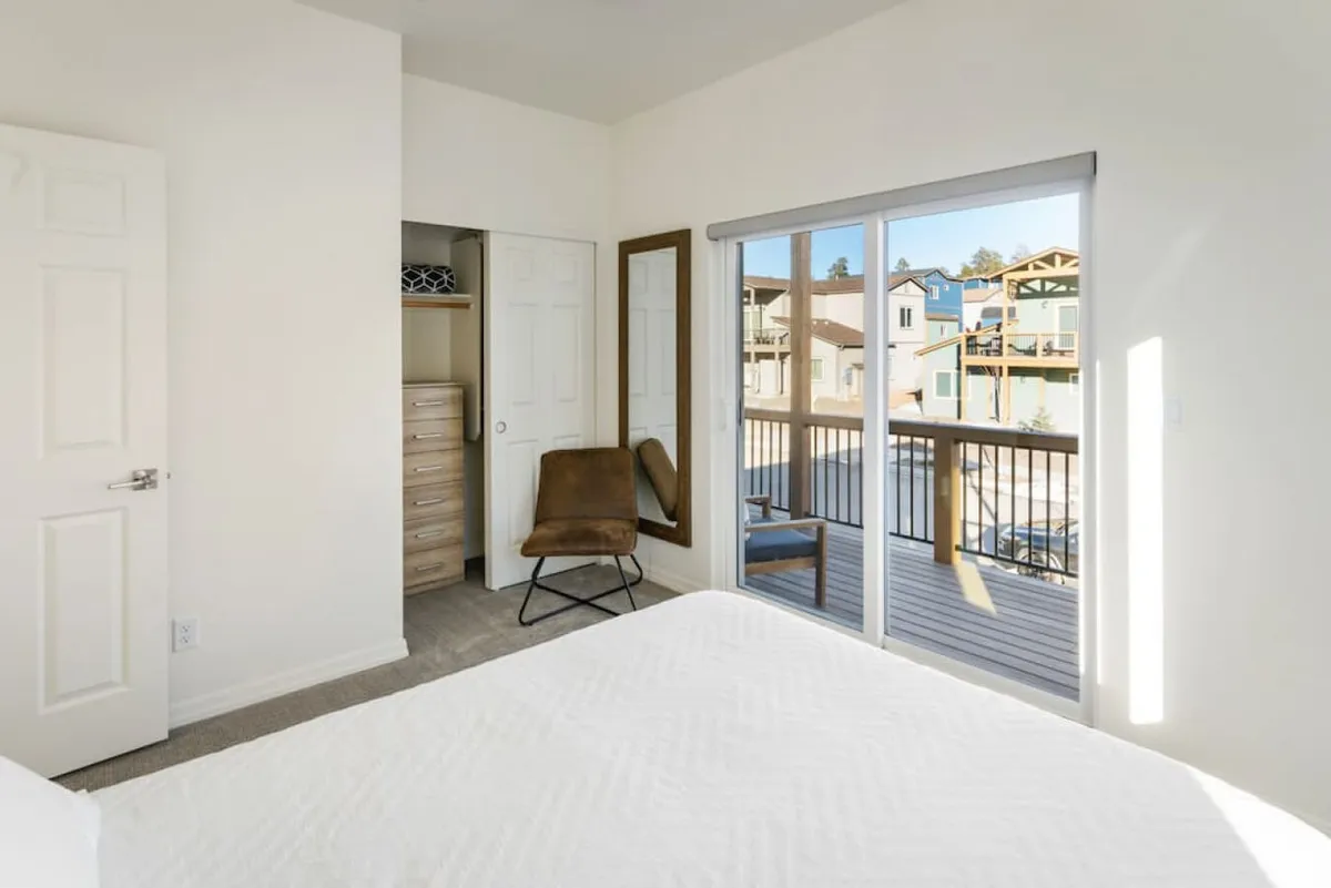 A calm, sunlit bedroom featuring a bed positioned near a sliding glass door. The door opens to outdoor views, and a cozy chair sits nearby, inviting you to enjoy the sunlight and neighborhood scenery. Soft morning light pours in, emphasizing the tranquil ambiance and the connection to the outdoors.