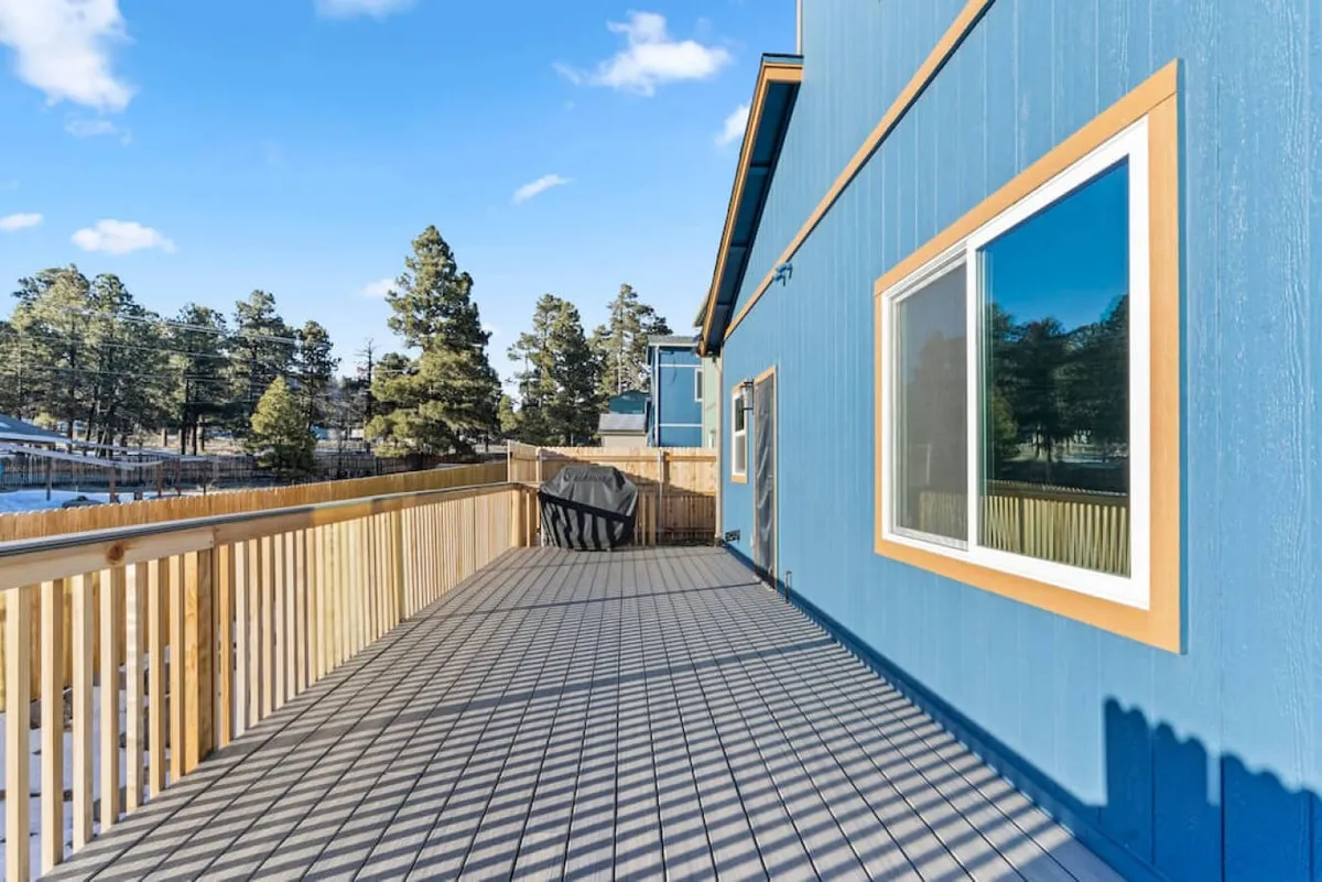 A view of a spacious deck attached to the blue house, bordered by lush surroundings. The deck is set up for gatherings, with space for barbecuing, relaxing, or simply enjoying the outdoors with family and friends.